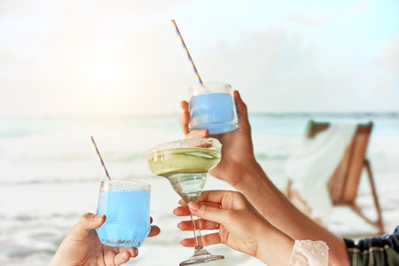 Beach party. Friends gathering, hands clinking glasses with refreshing cocktails against sea and beach background with sunlight. Concept of summer, vacation, travelling, relaxation, natureの写真素材