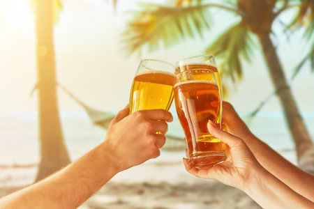 Human hand holding glasses with chill frothy drink, lager beer against sunny tropical background with palms, ocean and hammock. Concept of summer, vacation, travelling, relaxation, natureの写真素材