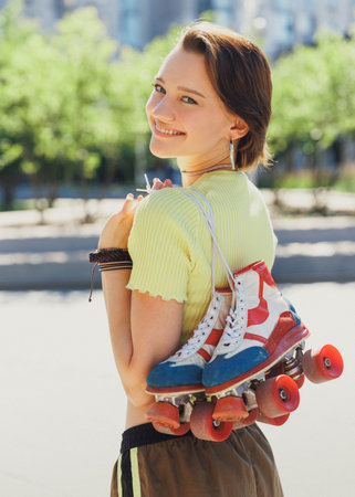 Portrait of young attractive teen girl posing holding vintage roller skaters, looking over shoulder against blurred background of park view. Concept of sport, active lifestyle, youth, hobby.の写真素材