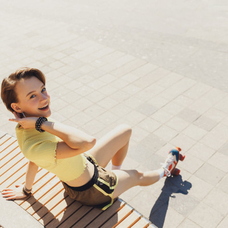 Aerial view. Portrait of young attractive girl, teenager wearing sporty clothes and retro roller skaters sitting on bench in sunny day at park. Concept of sport, active lifestyle, youth, hobby.の写真素材