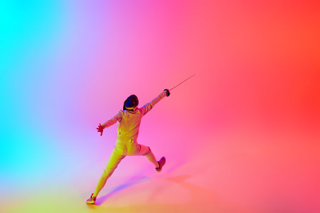 Fencer in gear and mask with their rapier extended forward, performing dynamic leap in mixed neon lights against gradient studio background. Concept of professional sport, competition, achievement. Adの写真素材