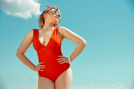 Joyful woman in red swimsuit and white cat-eye sunglasses, striking pose under sunny sky. Vintage summer charm. Concept of hot summer holiday, weekends, vacation, relaxation.の写真素材