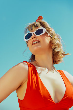 Happy woman in vibrant red swimsuit and white sunglasses, standing against bright blue sky. Retro...