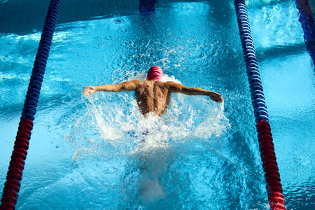 Swimmer, seen from above, powers through water with butterfly stroke, his muscular back glistening...