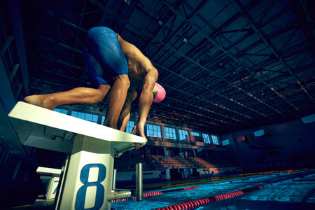 Ready to dive. Low angle view of young muscular man, swimmer, wearing goggles and cap, in low position on starting block in swimming pool. Concept of aquatic sport, preparation to competition, energy.の写真素材