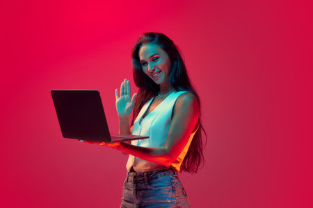 Young successful woman working remotely and waving hello holding meeting online in neon colors against magenta studio background, Trendy colors. Concept of study and working online, technology.の写真素材