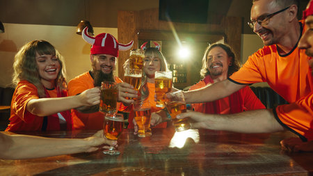 Friends, football fans in orange shirts raise their glasses in celebratory toast, their faces...