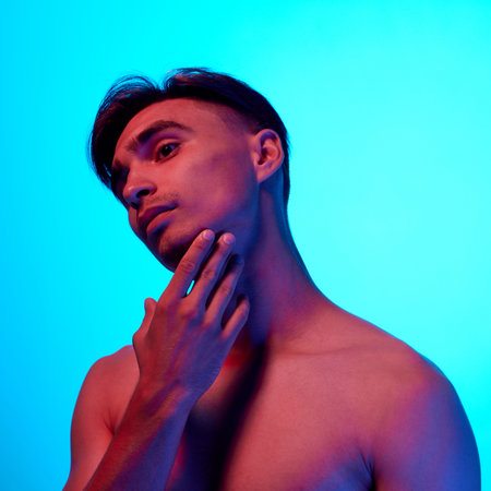 Portrait of shirtless brunette man posing gently touching his chin in neon light against blue gradient background. Concept of beauty, care, cosmetology, wellness, masculinity. Adの写真素材