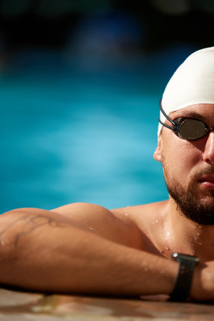 Cropped portrait of confident look of swimmer on edge of outdoor-pool. Athlete man wearing cap and...