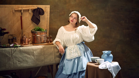 Young chubby woman, looks as maiden, in historical dress posing surrounded by vintage kitchenware and herbs. Concept of bar, pub, Bavarian style, Oktoberfest. Comparisons of eras.の写真素材