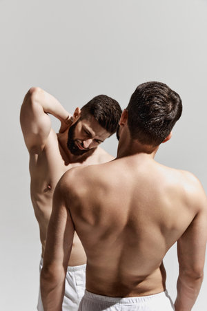 man in white shorts posing with pained expression, while another mans blurred silhouette frames scene against warm grey background. Concept of male health, beauty and fashion, masculinity. Ad.の写真素材