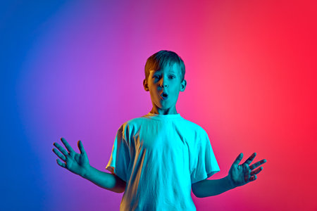 Surprised little boy raising hands of astonishment in neon light against gradient blue-pink studio background. Concept of children, human emotions, fashion and style, lifestyle. Adの写真素材