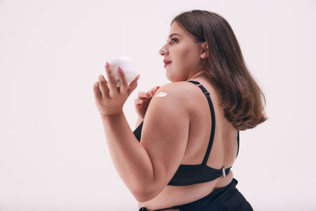 Rear view photo of chubby, young woman posing in black swimwear and applying cream to protect her...
