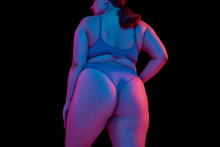 Cropped rear view of lower body, buttocks of plus size model in lingerie in neon light against black...