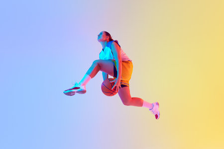 Photo of young woman, basketball player leaping in mid-air to make powerful slam dunk in neon light...