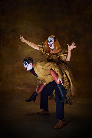 Woman in clown make-up, sitting on partners back, both dressed in vintage attire, striking playful...