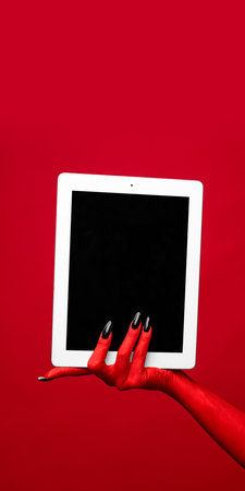Horror red devils hand with blank long nails holding tablet with blank screen against red studio background. Concept of season sale, promotion, celebration, web. Negative space to insert text. Adの写真素材