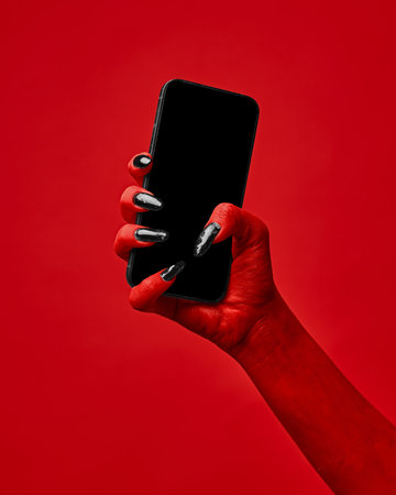 Horror red devils hand with blank long nails holding smartphone with blank screen against red studio background. Concept of season sale, promotion, celebration, web. Negative space to insert text. Adの写真素材
