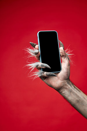 Werewolfs hand with long fingers and nails, paw holding modern smartphone with blank screen against...