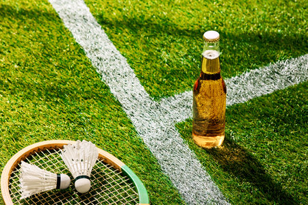 Badminton racket and shuttlecocks on green turf beside cold beer glass bottle. Perfect blend of sport and relaxation on sunny day, captured on field. Concept sport, blend of active and leisure timeの写真素材