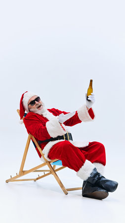 Santa leans back in chair, enjoying beer in his hand, wearing sunglasses, looking relaxed and carefree against white background. Concept of winter holidays, Holly Jolly mood, celebration. Adの写真素材