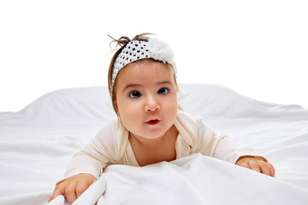 Curious tot in cute outfit peeks over blanket on soft bed with white bedding against studio background. Concept of beauty, parenthood, childhood, life and birth.の写真素材