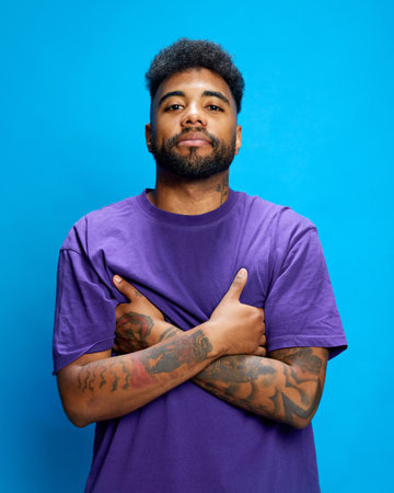 Young man in purple shirt stands confidently, crossing his tattooed arms, looking at camera with confidence against blue background. Concept of human emotions, youth, social media, fashion and style.の写真素材