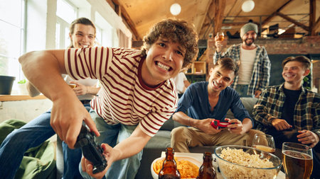Leaning in close, one player has big grin while gripping controller. Groups energy high, snacks and drinks scattered on table. Concept of football fans, sport evens, video games and cybersport.の写真素材