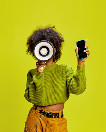 Woman with megaphone in one hand and smartphone in other, ready for social media against green studio background. Concept of announcement, communication, digital marketing, public interaction.の写真素材