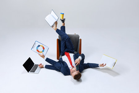Businessman lying on ground, his lags in multiple directions, managing papers, laptop, and phone calls. Chaos meets productivity. Concept of business, work overload, multitasking, corporate stressの写真素材
