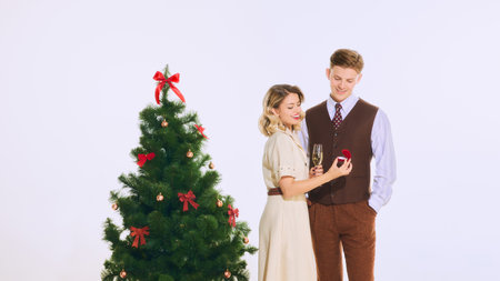 Woman admires ring in box while man smiles warmly, standing near festive Christmas tree with holiday...