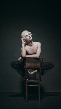 Cross-legged and thoughtful, young man sits elegantly on chair, aristocratic presentation, framed by classic postures in minimalistic style. Concept of fusion and modern, art, music and party, ballet.の写真素材