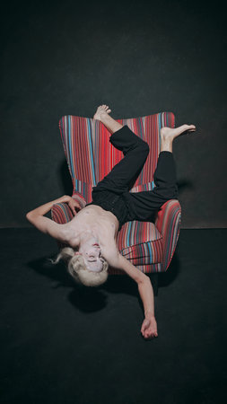 Young man lying on armchair in upside-down pose against vintage style studio background. Blurring...