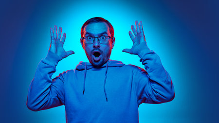 Young man in glasses raising hands with shocked, surprised expression in neon light against gradient studio background. Concept of human emotions, beauty and fashion, wow.の写真素材