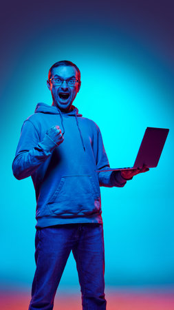 Football fan, young man dressed casually, holding laptop and clenching hand neon light against gradient background, as his favorite sport team win. Concept of human emotions, beauty and fashion. Adの写真素材