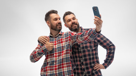 Two young guys, twin brothers dressed matching checked shirts and jeans, hugs and taking selfie...