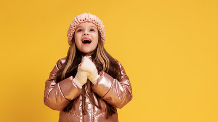 Excited little girl, dressed warm winter clothes, looking up and holds hands together, as she see a wonder against yellow studio background. Concept of childhood, lifestyle, expressions, trendy.の写真素材