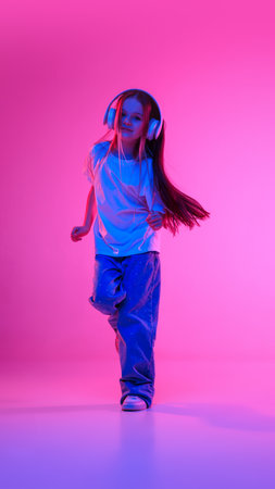 Dynamic shot of little charming girl dancing with joy while listening music in headphones in neon light against vivid gradient background. Concept of childhood, lifestyle, expressions, trendy. Adの写真素材