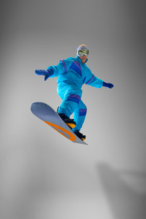Athletic snowboarder balancing gracefully in mid-air jump, dressed vibrant blue jumpsuit against grey studio background. Excitement of snowboarding. Concept of extreme sport, energy, speed, fashion.の写真素材