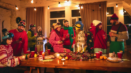 Cozy party unfolds as friends in colorful winter attire toast around wooden table with lit candles, holiday pastries, fresh oranges, and cheerful decorations. Concept of holidays, Christmas party. Adの写真素材