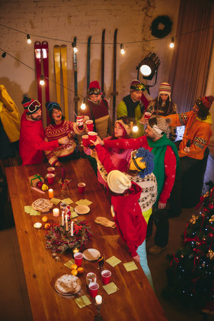 Friends in cozy winter-themed outfits toast with red cups, gathered around table decorated with wreaths, oranges, and candles, enjoying holiday festivities. Concept of holidays, Christmas party.の写真素材