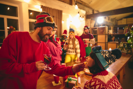 Cheerful group in winter sweaters gathers around rustic table laughing in festive mood. Couple...
