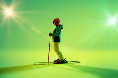 Female skier stands poised, dressed in colorful winter gear against radiant green gradient background, her poles upright. Concept of extreme sport, holidays.の写真素材