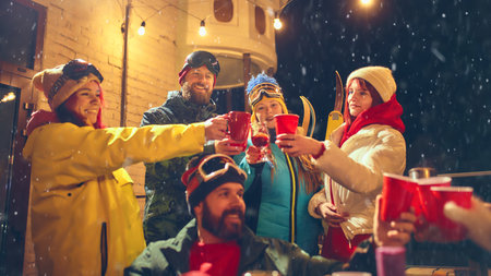 Snowy evening celebration with friends in vibrant ski outfits, raising red cups and enjoying festive...