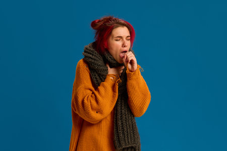 Portrait of very sick, ill young woman wrapped in scarf, dressed casually, and cough against dark...