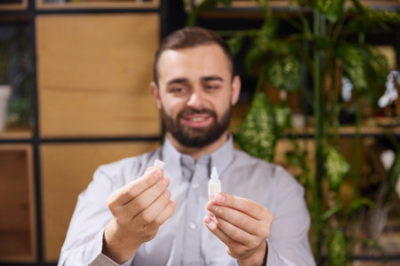 Young man opens bottle of eye drops and prepare to use them while sitting at desk in his workplace. Diseases and treatments. Concept of illness, health insurance, pharmaceutical, medicine. Ad.の写真素材