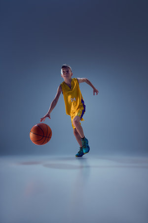 Dynamic photo of junior basketball athlete, running with ball, ready to perform a slam dunk against grey studio background. Concept of sport, leisure activity, hobby, competition.の写真素材