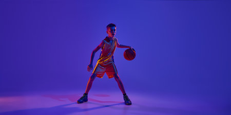 Confident, teenage ball stands ready to start game and get victory in this decisive basketball match in neon light against gradient studio background. Concept of sport, leisure activity, competition.の写真素材