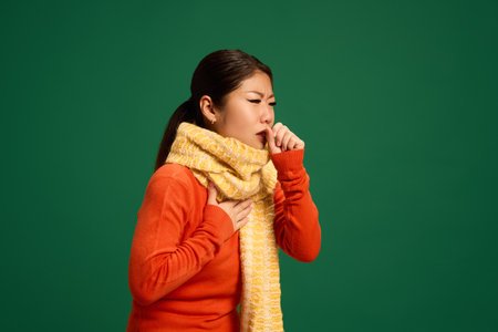 Woman coughing into hand, wearing scarf, showing symptoms of viral respiratory illness, bronchitis, or flu causing persistent cough. Concept of healthcare, medicine, infection and treatment. Ad.の写真素材
