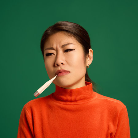 portrait of Asian woman holding thermometer in mouth, showing signs of fever or viral illness,...